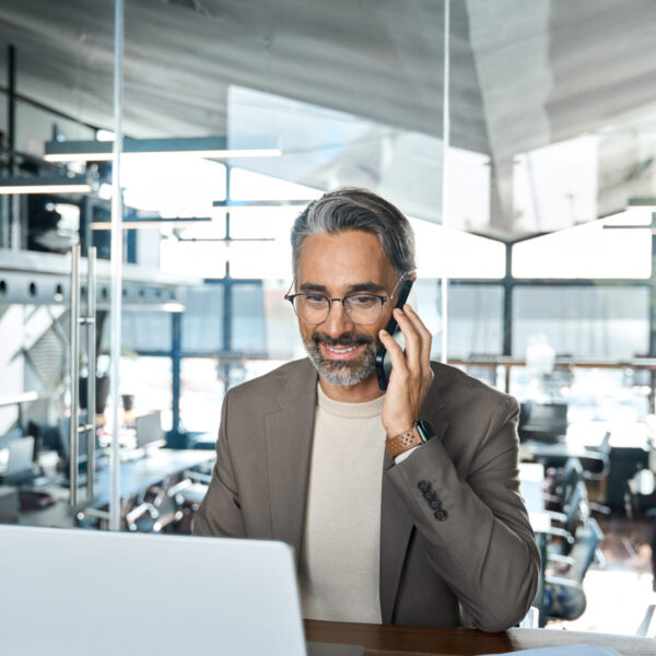 Happy mid aged business man ceo talking on cell phone working on laptop in office. Busy smiling mature professional businessman executive making call on cell using computer sitting at desk. Vertical